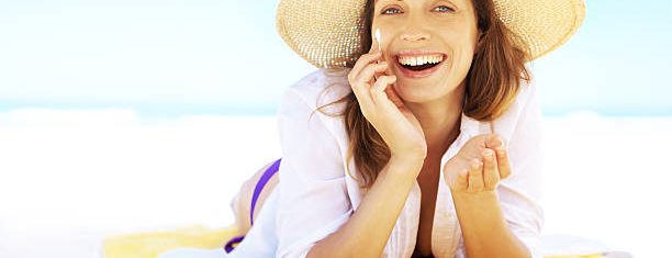 Mulher sorrindo com chapéu de Sol passando protetor solar na praia | Mulher sorrindo e brincando com cabelo na praia | Cuidados com pele para o verão